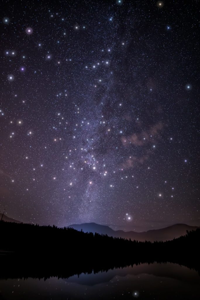 Starry night above dark mountains & trees. Their reflection shimmers in a tranquil lake, evoking contemplation.